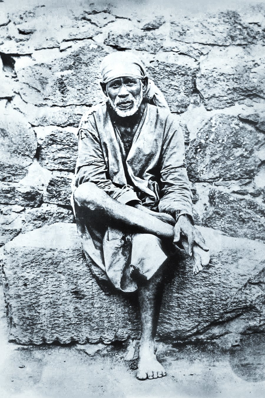 Original photograph of Shirdi Sai Baba, seated on a stone before a stone wall
